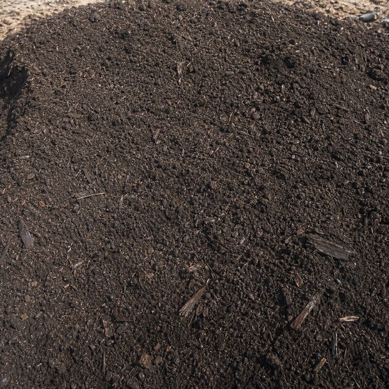 Compost maduro: material marrón oscuro, fino y homogéneo, con aspecto de tierra húmeda y algunas astillas gruesas sin degradar.