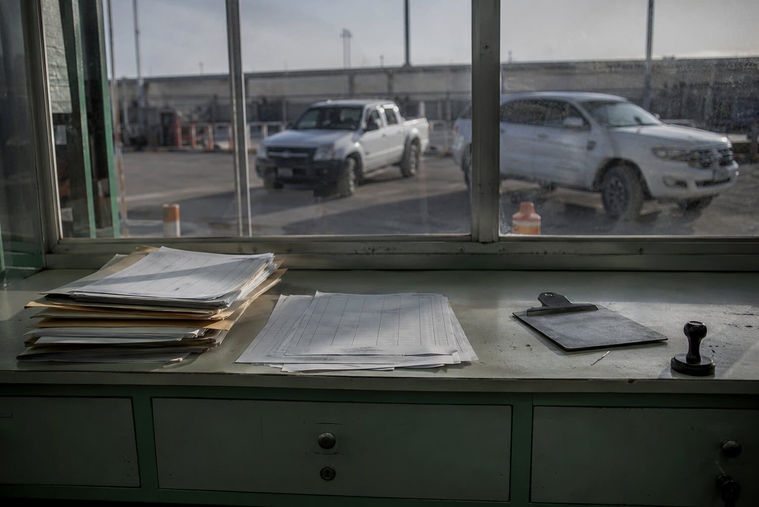 Mostrador de una planta de verificación con una pila de carpetas, planillas, un portapapeles y un sello; por el ventanal se ven dos camionetas de trabajo esperando turno.