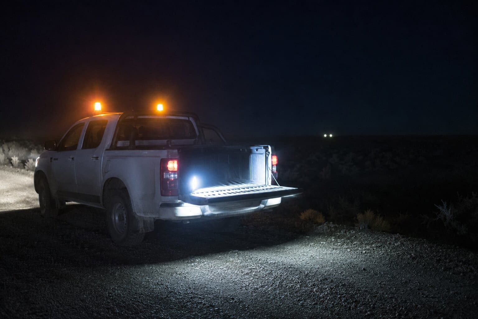 Camioneta de trabajo detenida de noche en una huella de ripio, con las balizas encendidas y un foco portátil iluminando el suelo desde la caja.
