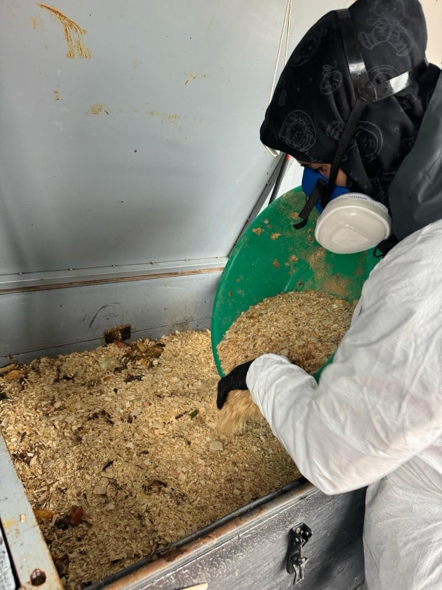 Un operario con mameluco y guantes vuelca material estructurante desde un balde dentro de una compostera.