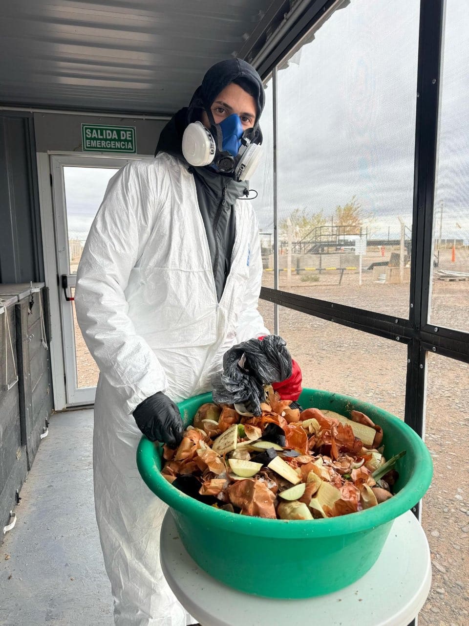 Un operario con mameluco blanco, guantes y respirador sostiene una palangana con residuos orgánicos de cocina dentro del módulo de compostaje.